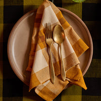 A styled tabletop setting featuring a folded ochre yellow and cream plaid napkin on a beige ceramic plate, with gold-toned cutlery. The setting is placed on a dark green and black buffalo plaid tablecloth, with a green glass mug visible in the background.