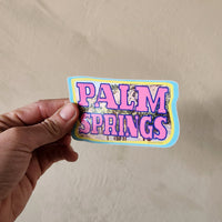 Hand holding a 'Palm Springs' sticker against a beige background