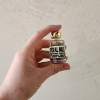 A hand holding a small glass bottle of Feral Milk Functional Fragrance with a gold cap against a textured off-white wall.