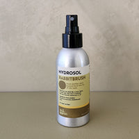 Hydrosol Rabbitbrush room spray in a silver aluminum bottle with a black spray nozzle and a beige and brown label, set against a textured beige background.
