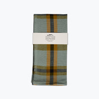 A folded cotton napkin with a muted sage green and mustard yellow plaid pattern, wrapped in a white paper band labeled 'ALL Roads COTTON NAPKINS'.