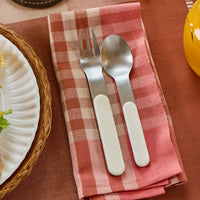 Silver fork and spoon on a pink checkered napkin with a plate and cup in the background.