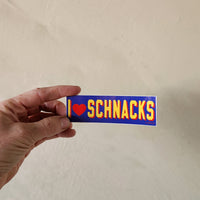 Hand holding a rectangular sticker with 'I ❤️ Schnacks' text on a beige background
