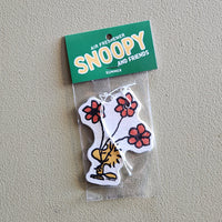 A Snoopy and Friends air freshener sticker in its clear plastic packaging with a green header card, placed on a textured beige wall. The sticker features a white dog body with red flowers and a yellow bird on it.