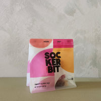 A bag of Sockerbit Sweet Gummy Mix standing upright on a beige surface against a textured off-white wall. The packaging features a bold, modern design with pink, orange, and yellow geometric shapes and black text.