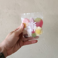 A hand holding a clear plastic bag of Swedish candy SOCKER BIT against a textured beige wall.