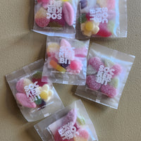 Six clear plastic bags of Sockerbit sour candy arranged on a textured beige surface. The candy inside is various colors including pink, yellow, and green, and the packaging features the Sockerbit logo.