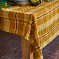 A mustard yellow and cream plaid tablecloth draped over a wooden table, styled with plates, a matching napkin, and greenery on a brick patio.
