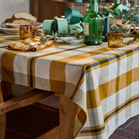 A rustic wooden dining table set with a yellow, white, and blue plaid tablecloth, amber glass tumblers, green glass bottles, and fresh herbs in a warm, styled dining room setting.