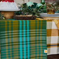 A green and teal plaid table runner with fringe is draped over a table, alongside a yellow-and-white plaid cloth. The scene is styled with dishes, a cake, greenery, and a bottle in a rustic indoor setting.