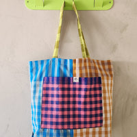 Colorful checkered tote bag with a pink pocket hanging on a green hook against a beige wall.