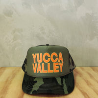 Army green camo foam trucker hat with the text Yucca Valley.