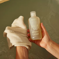 Hand holding a bottle of 'Desert Cedar' body wash with a washcloth against a green background