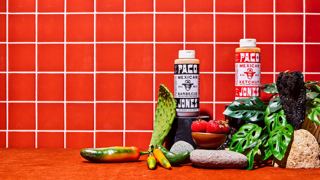 Two bottles of PACO Mexican Ketchup and BBQ Sauce displayed on a wooden surface with various Mexican ingredients, including a jalapeño, a prickly pear cactus, strawberries, and green leaves, against a red tiled background.