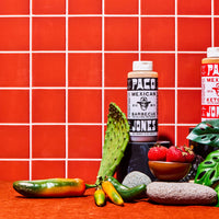 Two bottles of PACO Mexican Ketchup and BBQ Sauce displayed on a wooden surface with various Mexican ingredients, including a jalapeño, a prickly pear cactus, strawberries, and green leaves, against a red tiled background.