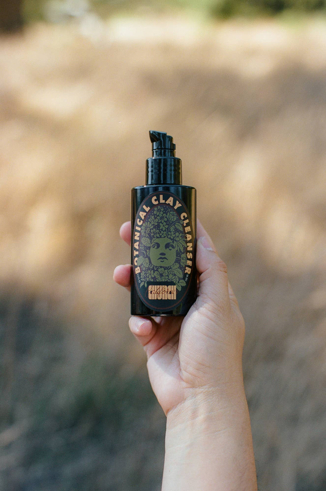 A hand holding a black pump bottle of Botanical Clay Cleanser against a blurred outdoor background.
