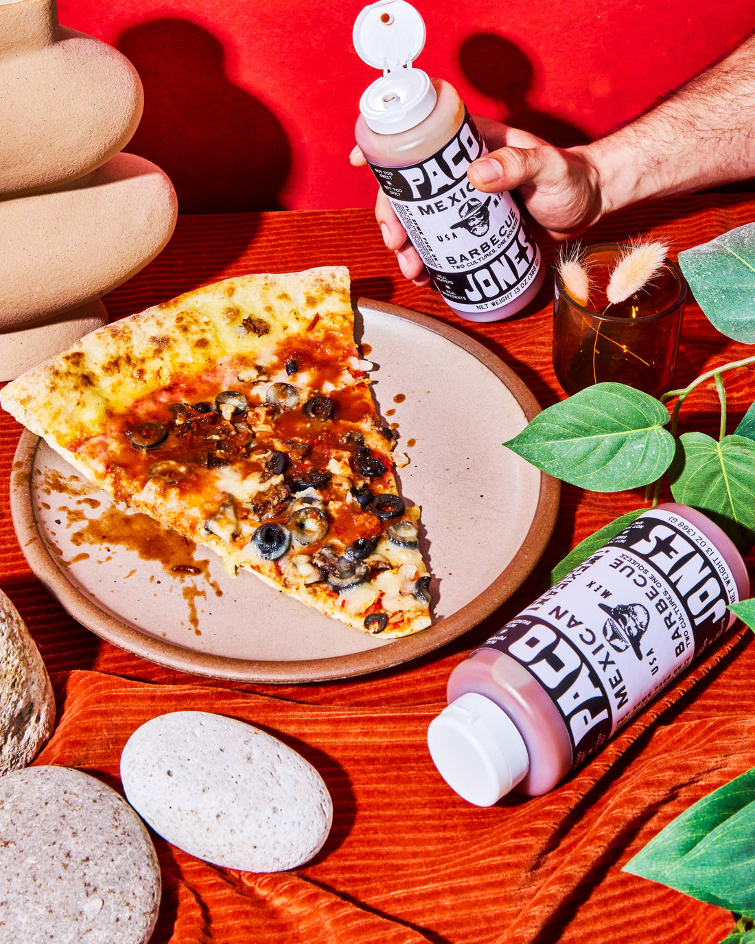 A hand holds a squeeze bottle of Mexican Barbecue sauce next to a slice of pizza on a plate, with another bottle lying on an orange textured cloth and more food props around it.