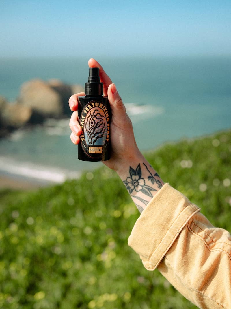 A hand holding a black spray bottlE against a blurred outdoor background of green grass and the ocean.