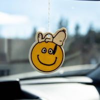 Smiley face air freshener hanging from a car's rearview mirror with a blurred outdoor background.