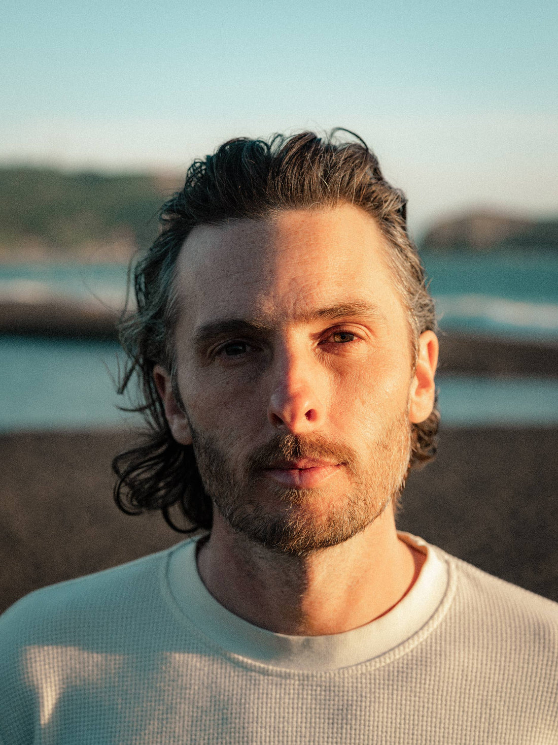 A close-up portrait of a man with wavy dark hair and a short beard, wearing a light-colored waffle-knit crewneck shirt, set against a blurred beach background at sunset.