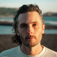 A close-up portrait of a man with wavy dark hair and a short beard, wearing a light-colored waffle-knit crewneck shirt, set against a blurred beach background at sunset.