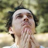 A close-up outdoor portrait of a man with dark hair and a mustache, looking upward with his hands clasped together in a prayer-like pose against a blurred green and beige background.