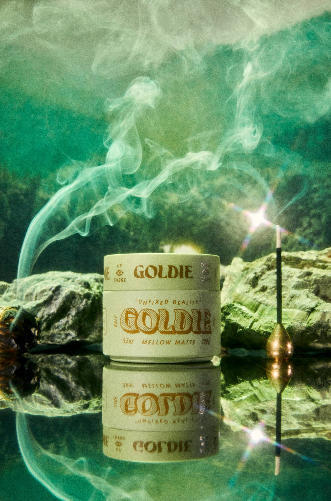 A jar of Goldie Mellow Matte sits on a reflective surface with a smoking incense stick and green mist in the background.