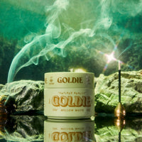 A jar of Goldie Mellow Matte sits on a reflective surface with a smoking incense stick and green mist in the background.
