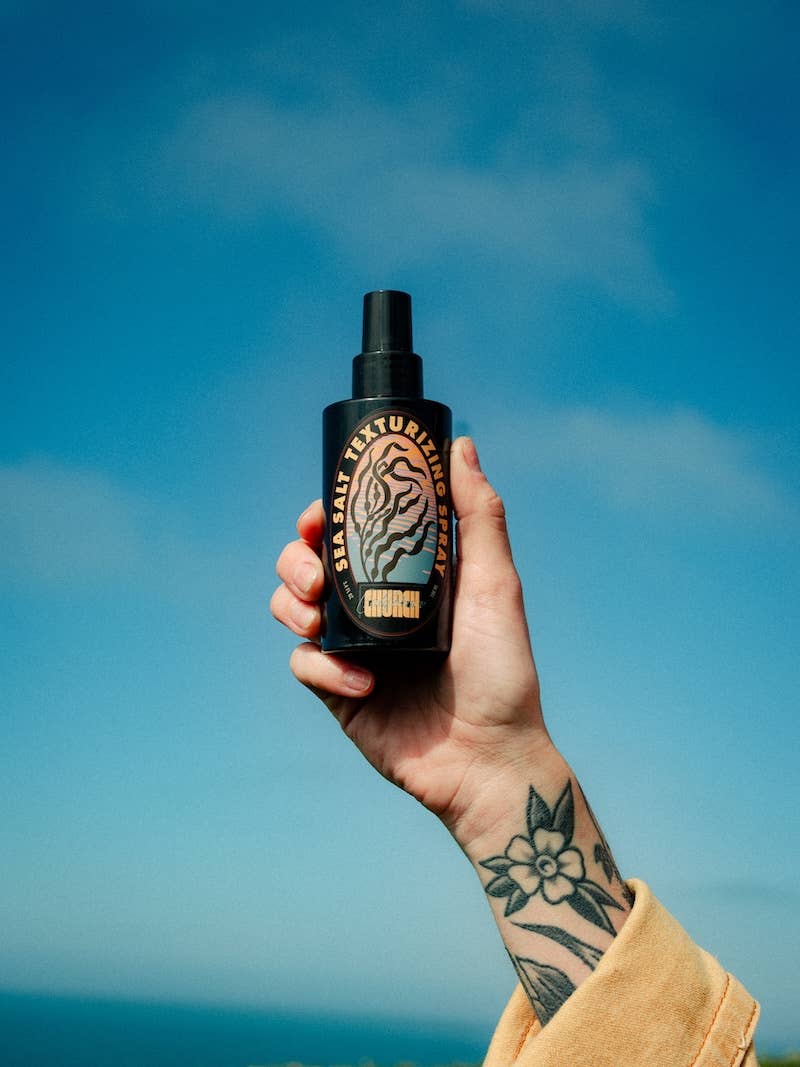 A hand with a floral tattoo holds up a black spray bottle of hair texture spray against a bright blue sky background. The bottle features a black label with a stylized tiger graphic and gold text reading &