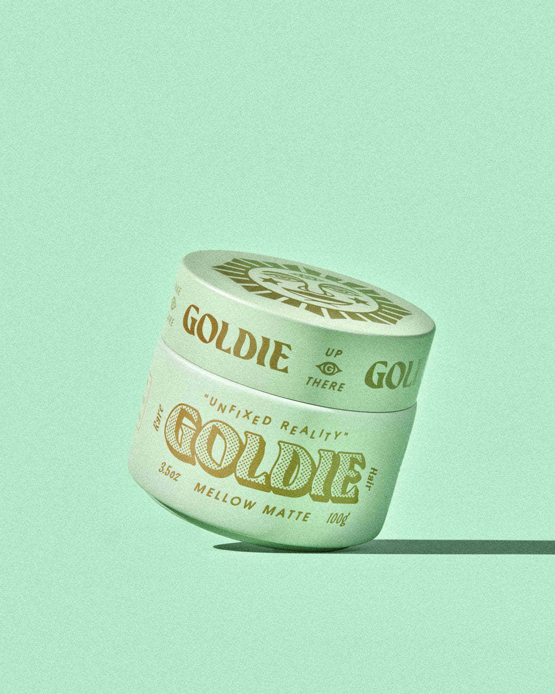 A single jar of Goldie Mellow Matte hair wax sits at an angle on a solid mint green background. The jar is a pale mint green with gold lettering and a matching gold lid, casting a soft shadow to the right.