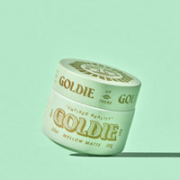 A single jar of Goldie Mellow Matte hair wax sits at an angle on a solid mint green background. The jar is a pale mint green with gold lettering and a matching gold lid, casting a soft shadow to the right.