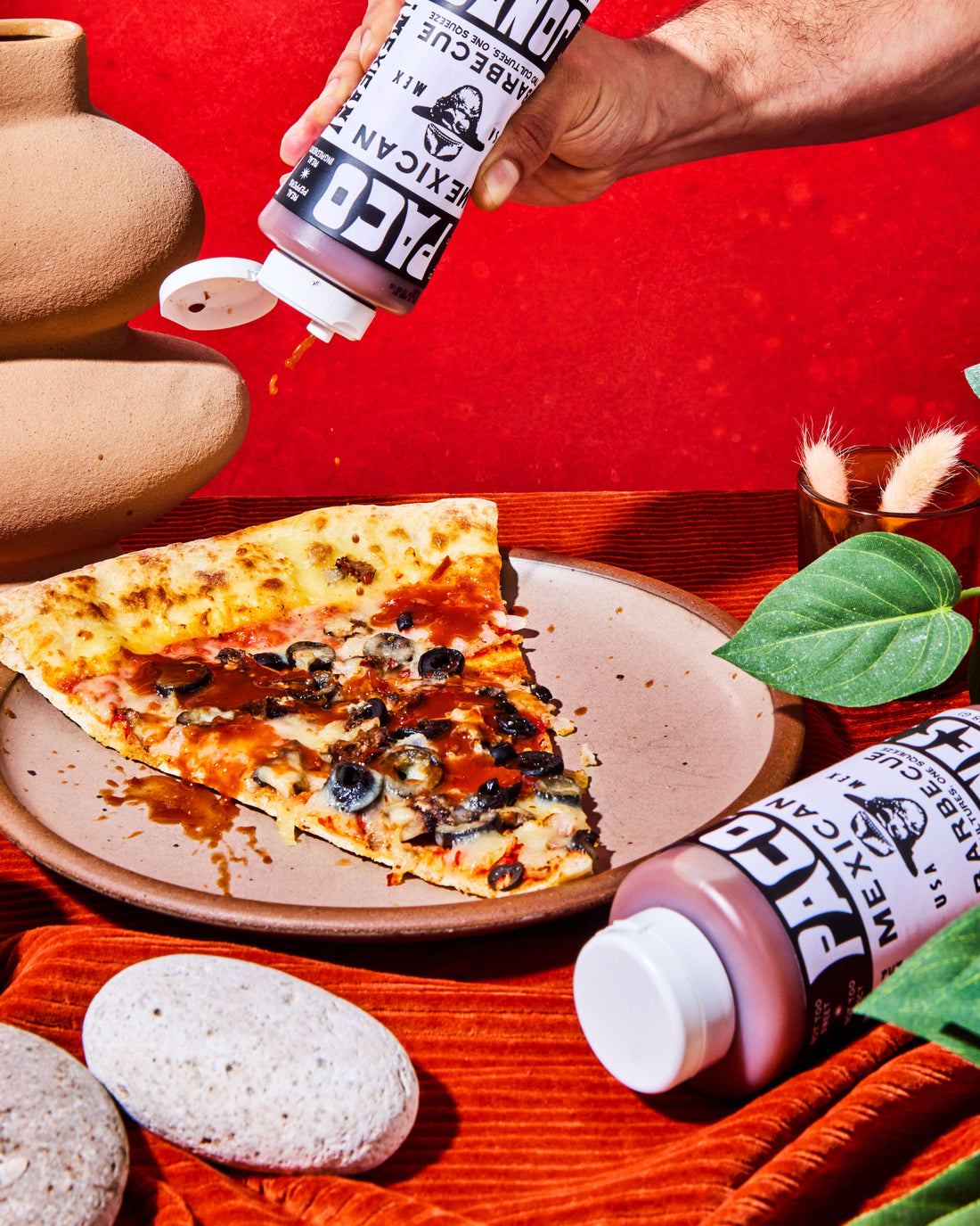 A hand squeezes a bottle onto a slice of pizza on a plate, with another bottle lying nearby on a rustic red and orange styled table.