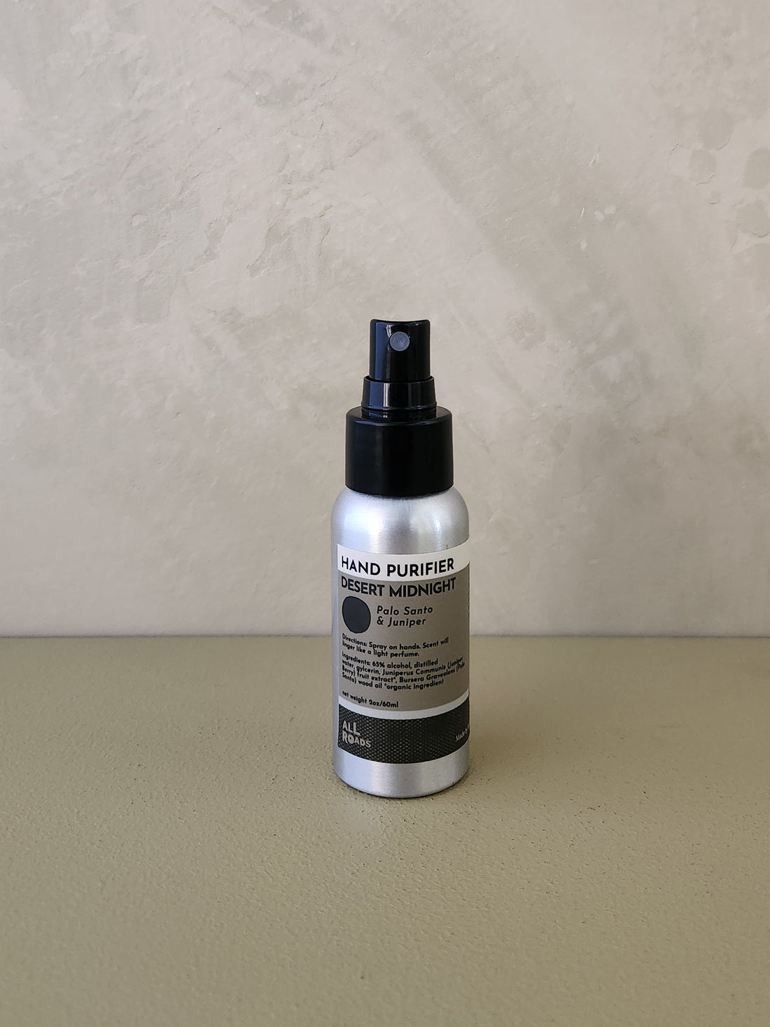 A silver spray bottle of Desert Midnight Hand Purifier with a black pump, standing on a beige surface against a textured beige wall.