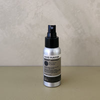 A silver spray bottle of Desert Midnight Hand Purifier with a black pump, standing on a beige surface against a textured beige wall.