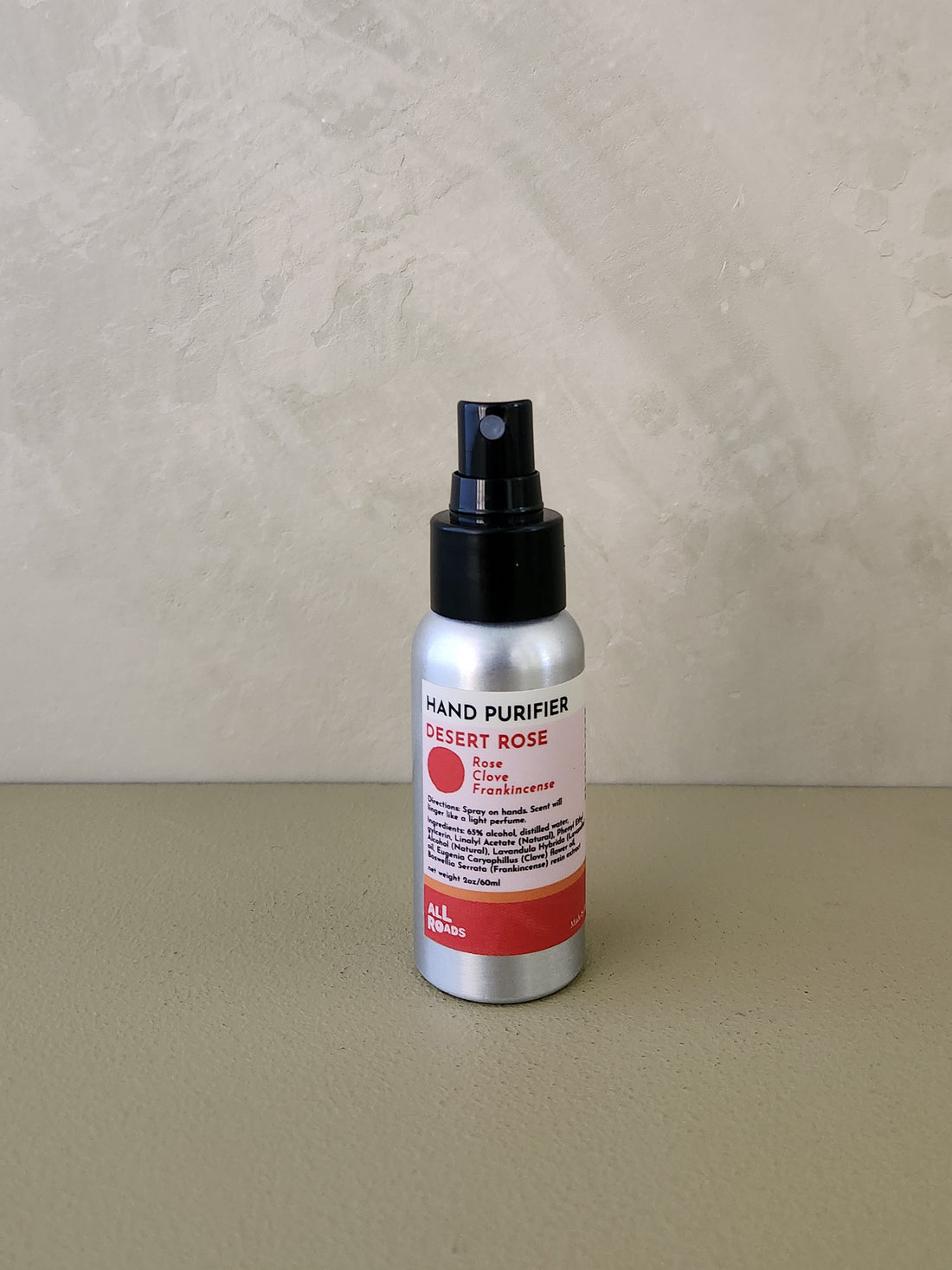A silver spray bottle of Hand Purifier Desert Rose hand cleanser with a black pump, standing on a neutral beige surface against a textured beige wall.