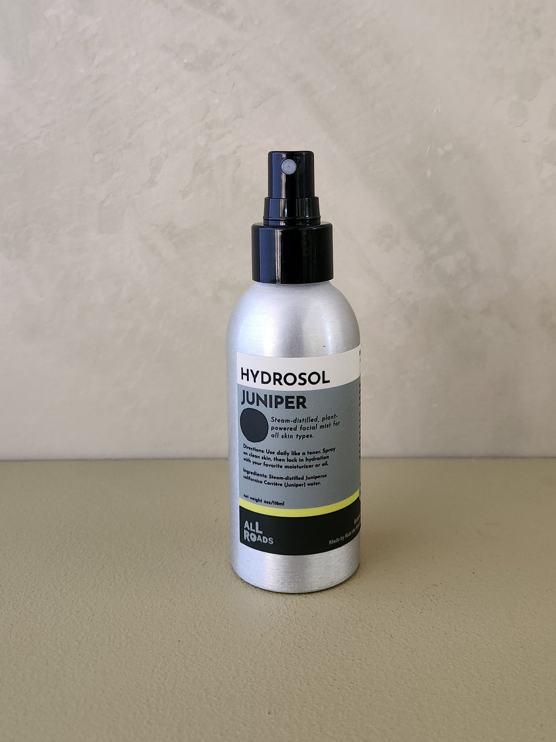 A bottle of Hydrosol Juniper room spray with a black spray top and a silver bottle body, standing on a beige surface against a textured beige wall.