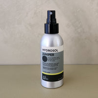 A bottle of Hydrosol Juniper room spray with a black spray top and a silver bottle body, standing on a beige surface against a textured beige wall.
