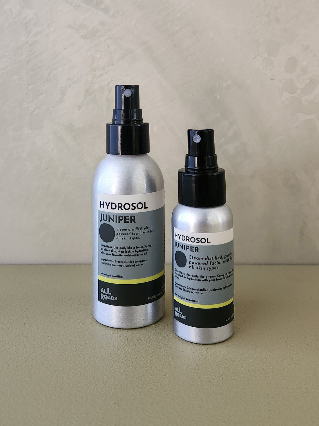 Two silver spray bottles of Hydrosol Juniper by All Spas on a beige surface against a textured beige wall.