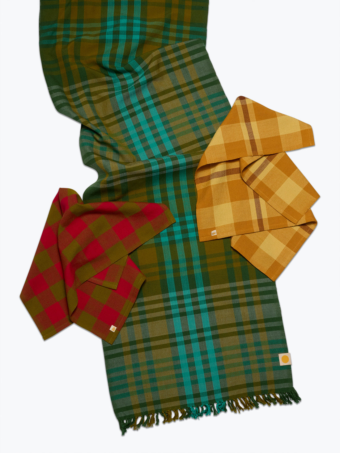 Three plaid tablelinens in different colorways (green/teal, mustard yellow, and red) arranged on a light gray background.