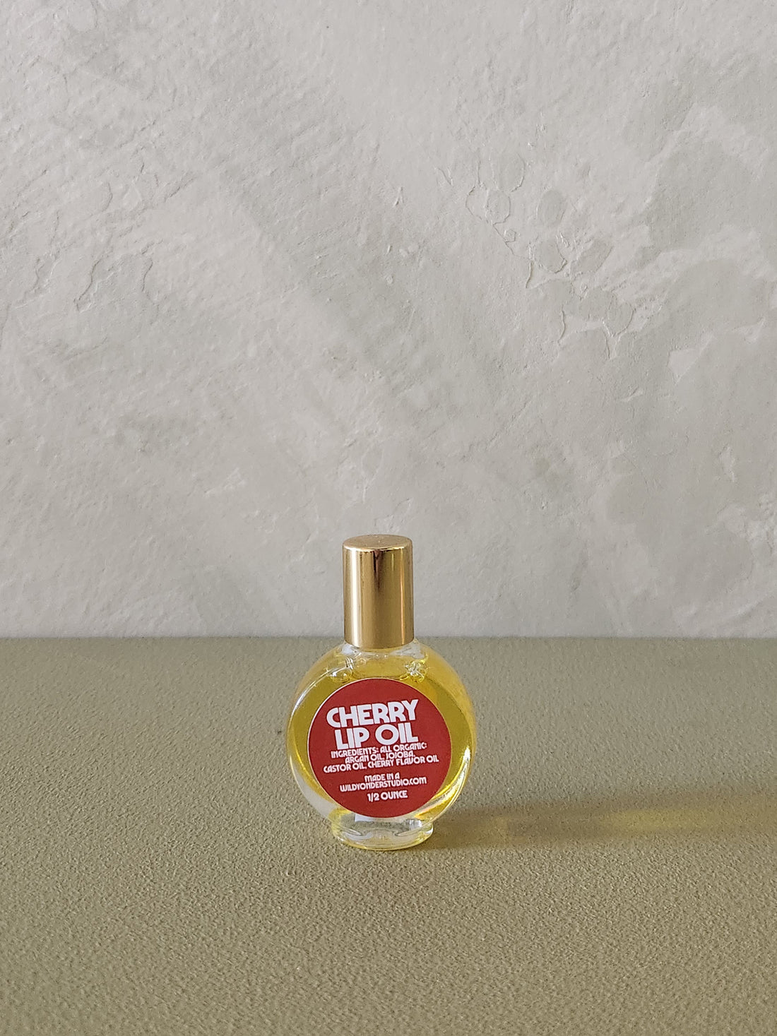 A small round glass bottle of cherry lip oil with a gold cap and a red label, placed on a textured beige surface against a light gray wall.