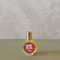 A small round glass bottle of cherry lip oil with a gold cap and a red label, placed on a textured beige surface against a light gray wall.