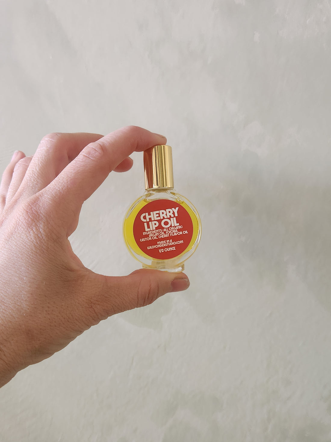 A hand holding a small round bottle of cherry lip oil with a gold cap against a textured off-white wall.