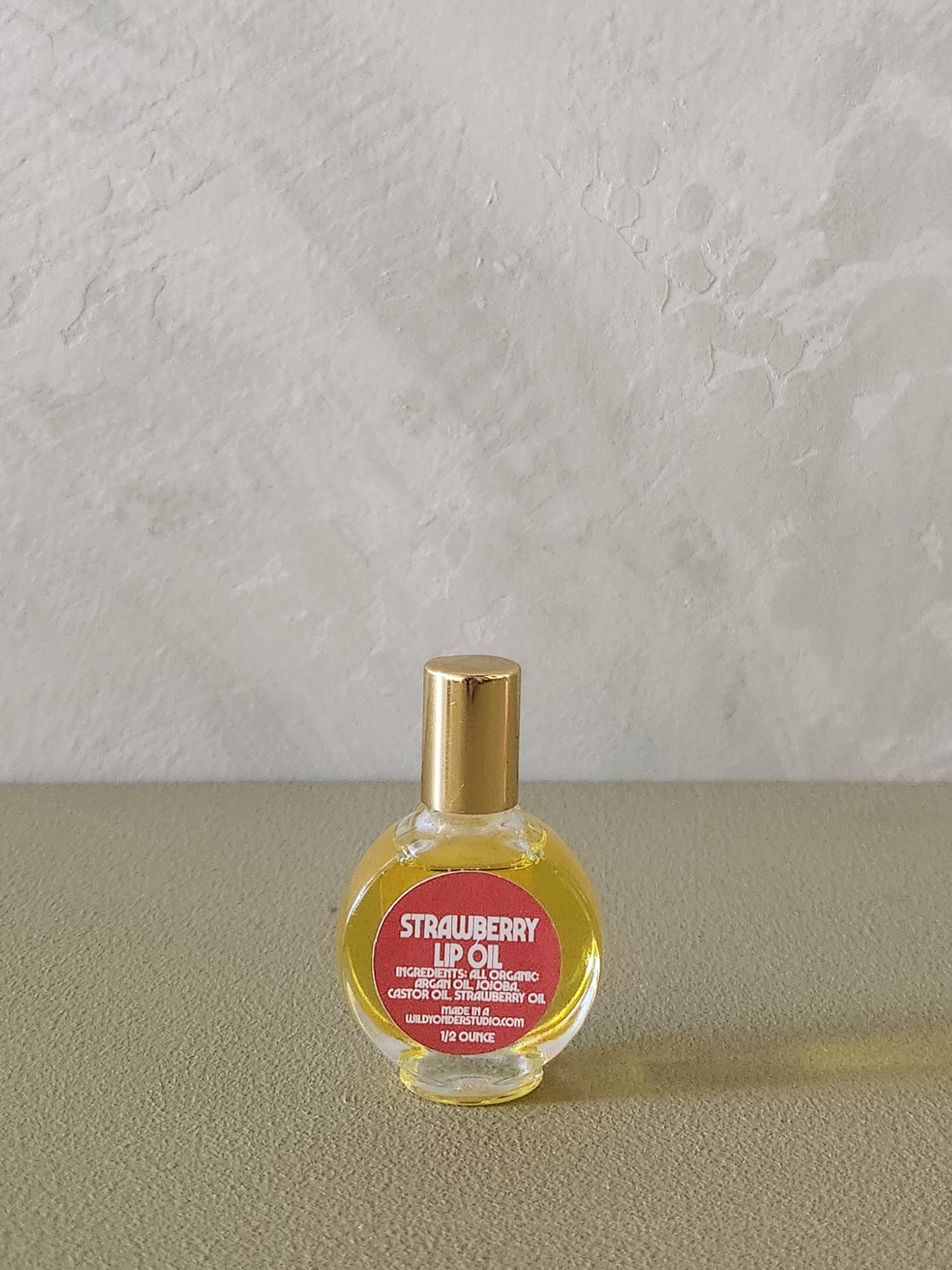 A small round glass bottle of strawberry lip oil with a gold cap and a red label, placed on a textured beige surface against a light gray wall.