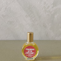 A small round glass bottle of strawberry lip oil with a gold cap and a red label, placed on a textured beige surface against a light gray wall.