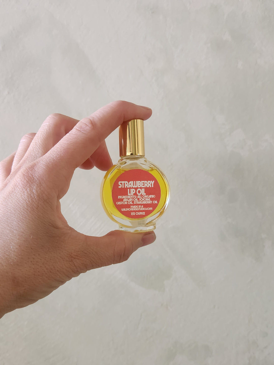 A hand holding a small round glass bottle of Strawberry Lip Oil with a gold cap against a textured light gray background.