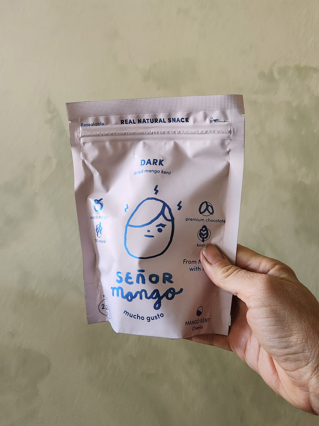 Hand holding a white snack package with blue text and branding against a plain background