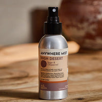 A 4oz spray bottle of 'Anywhere Mist' in the 'High Desert' scent, featuring a silver metallic bottle and black spray nozzle, set on a rustic wooden table with a warm, blurred background.