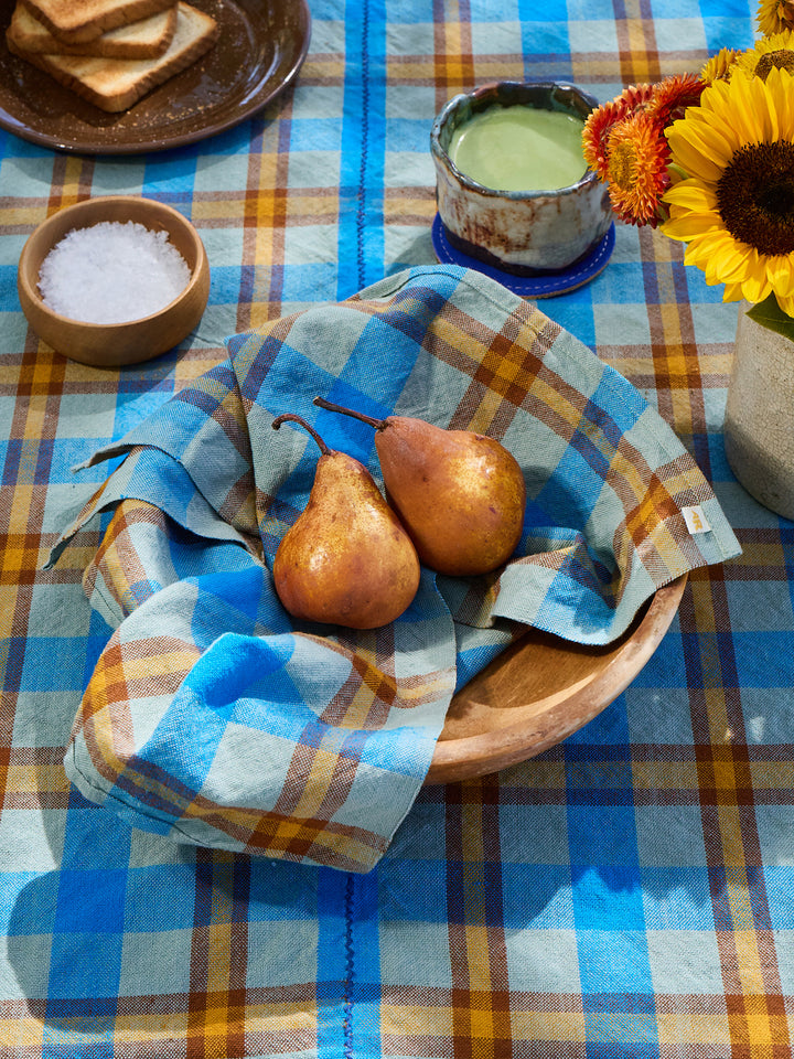Two pears in a wooden bowl on a checkered tablecloth with a plate of toast and a vase of sunflowers.
