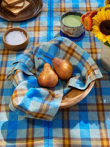 Two pears in a wooden bowl on a checkered tablecloth with a plate of toast and a vase of sunflowers.