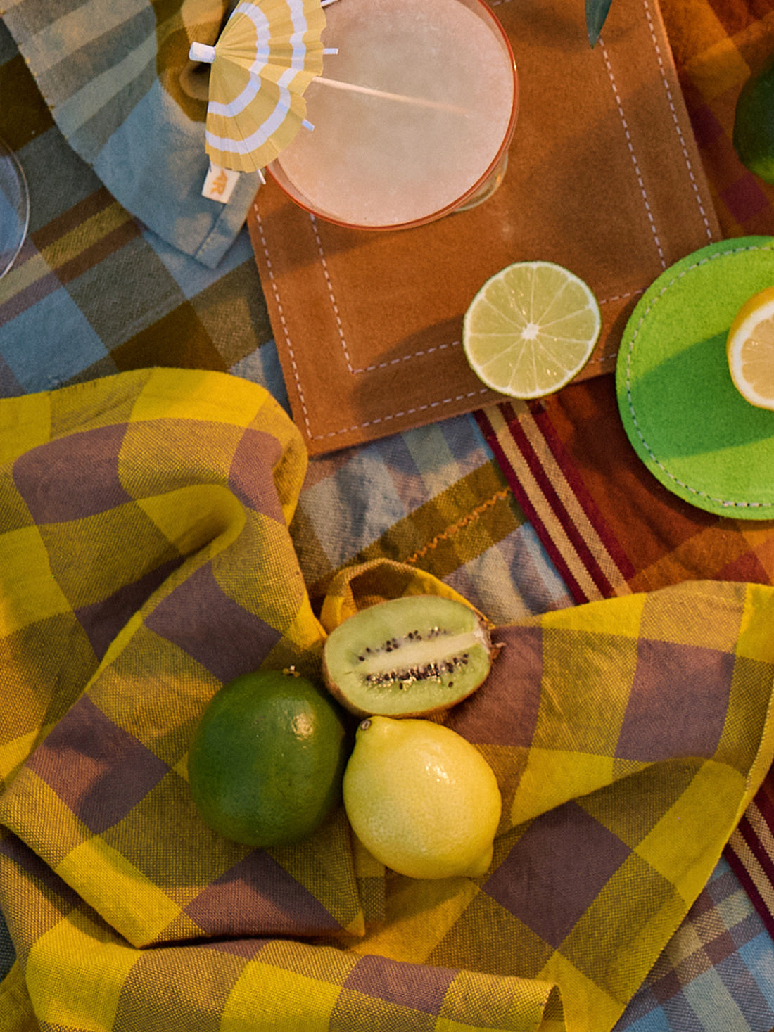 Fruits on a checkered tablecloth with colorful coasters and a drink.